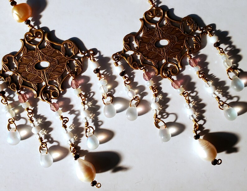 May include: A pair of gold-tone earrings with a delicate, ornate design. Each earring features a central pendant with a floral motif and a series of dangling beads, including white, pink, and iridescent beads.