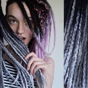 May include: A set of synthetic dreadlocks featuring a color gradient that transitions from deep black at the roots to a light grey at the tips. The texture is twisted and rope-like, with some strands showing a mix of black, grey, and light purple.