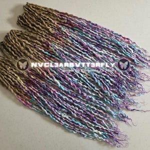 May include: Three bundles of handmade dreadlocks in shades of brown, purple, blue, and white. The dreadlocks are arranged diagonally across a light-colored surface. The text "NVCL3ARBVTT3RFLY designs by Cheyenne Le Hale" is visible.