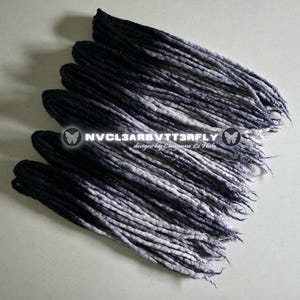 May include: A set of synthetic dreadlocks featuring an ombre color transition from solid black at the roots to a light grey at the tips. The text NVCL3ARBVTT3RFLY designs by Cheyenne Le Hale is overlaid in the center of the hair extensions.