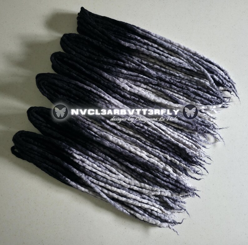 May include: A set of synthetic dreadlocks featuring an ombre color transition from solid black at the roots to a light grey at the tips. The text NVCL3ARBVTT3RFLY designs by Cheyenne Le Hale is overlaid in the center of the hair extensions.