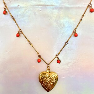 May include: A gold-toned metal necklace featuring a central heart-shaped locket with an embossed floral design. The chain is accented with small, evenly spaced, round orange beads that dangle from the links, creating a decorative pattern.