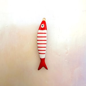 May include: A fish-shaped pendant featuring a glossy red and white striped pattern. The design includes a small circular eye detail near the top and a gold-toned metal loop at the head for attachment, creating a smooth, uniform finish throughout.
