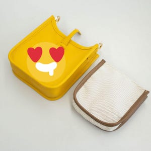 May include: A yellow handbag with a heart-eyes emoji design. The bag has a small handle and gold-colored hardware. A separate white pouch with a brown trim is next to the bag.