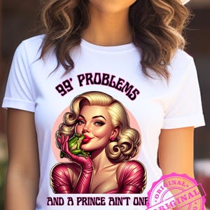 以下が含まれることがあります： 「99 Problems」の文字と、ブロンドの髪、赤い口紅、ピンクの手袋をした女性がカエルを持っているレトロなイラストが描かれた白いTシャツ。「And a Prince Ain't One」の文字がイラストの下にあります。ピンク色のスタンプには「Original」と書かれています。