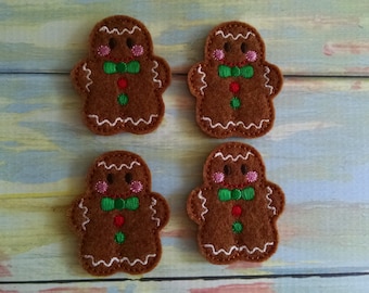 May include: Four brown felt gingerbread man ornaments. Each ornament has white icing details, pink cheeks, a green bow tie, and a red button. The gingerbread men are arranged on a light blue and yellow surface.