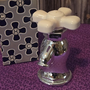 May include: A silver and white decorative bottle shaped like a faucet. The bottle has a white handle and a silver base. The bottle is sitting on a purple surface with a floral pattern.