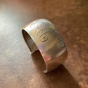 May include: A silver cuff bracelet with engraved initials. The bracelet has a curved, open design and a polished finish. The initials are in an elegant script, adding a personalized touch to the jewelry.