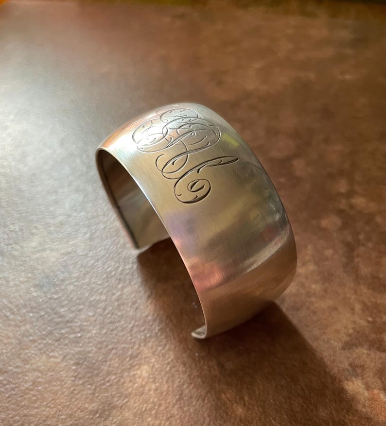 May include: A silver cuff bracelet with engraved initials. The bracelet has a curved, open design and a polished finish. The initials are in an elegant script, adding a personalized touch to the jewelry.