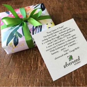 May include: A gift box wrapped in floral paper and tied with a bright green ribbon. A white card with black text is next to the box. The card reads "Rose Quartz & Pearls" and describes the bracelet.