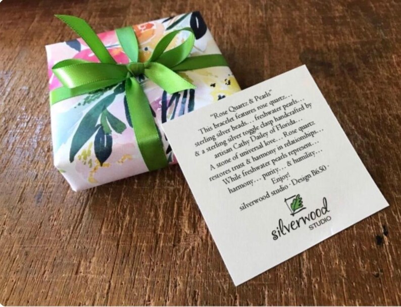 May include: A gift box wrapped in floral paper and tied with a bright green ribbon. A white card with black text is next to the box. The card reads "Rose Quartz & Pearls" and describes the bracelet.