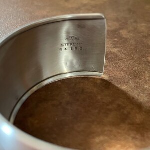 May include: Close-up of a silver-colored cuff bracelet. The inside of the bracelet is stamped with the word "STERLING" and the numbers "34 183". The bracelet has a smooth, polished finish.