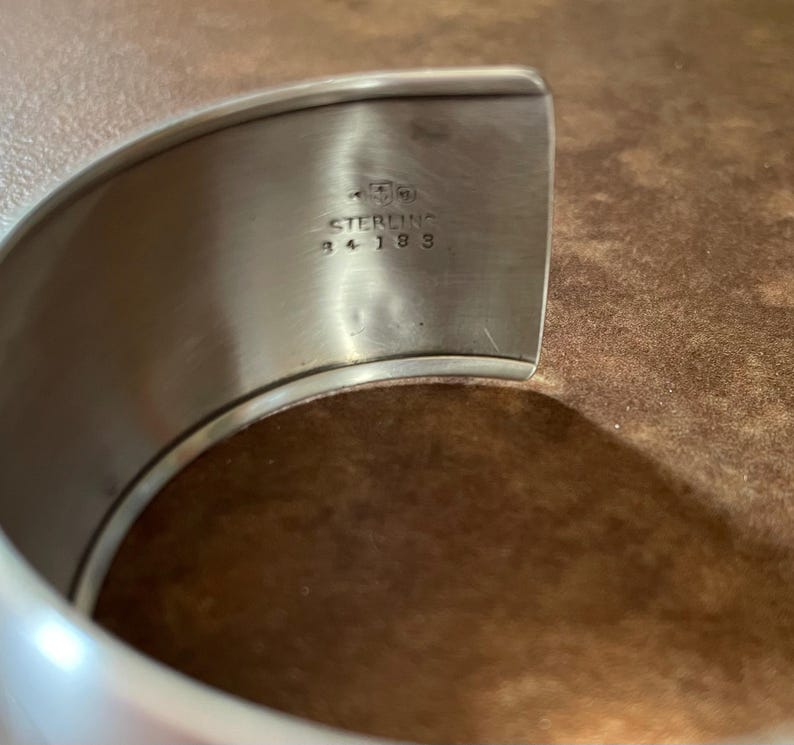 May include: Close-up of a silver-colored cuff bracelet. The inside of the bracelet is stamped with the word "STERLING" and the numbers "34 183". The bracelet has a smooth, polished finish.