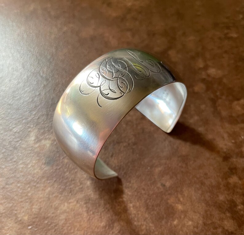 May include: A wide, silver cuff bracelet with an engraved monogram design. The polished metal reflects light, creating highlights and shadows. The open design allows for easy wear. A classic piece of jewelry.