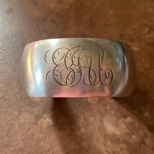 May include: A silver cuff bracelet with engraved initials. The wide band has a curved shape and a polished finish. The initials are in an elegant, flowing script. The bracelet is a classic piece of jewelry.