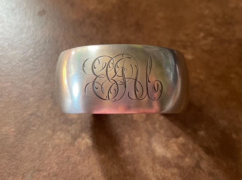 May include: A silver cuff bracelet with engraved initials. The wide band has a curved shape and a polished finish. The initials are in an elegant, flowing script. The bracelet is a classic piece of jewelry.