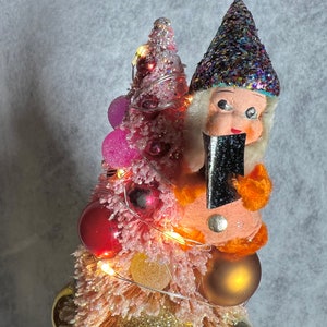 May include: A pink and gold miniature Christmas tree with a small gnome figurine holding a black gift. The tree is decorated with pink, red, and gold ornaments and has white lights.