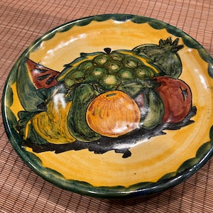 May include: A yellow ceramic plate with a green rim features a hand-painted design of a variety of fruits, including a watermelon, a green apple, a yellow pear, a red apple, and a green and yellow citrus fruit.