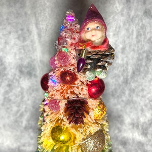 May include: A small, pink and yellow artificial Christmas tree decorated with red and gold ornaments, a pine cone, and a small elf figurine wearing a pink hat.