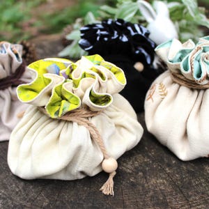 Puede incluir: Una colecci&oacute;n de bolsas de tela con cord&oacute;n en varios colores y patrones. Las bolsas est&aacute;n atadas con hilo y presentan bordes con volantes decorativos. Algunas tienen dise&ntilde;os florales o de helechos, y un detalle de cuentas de madera.