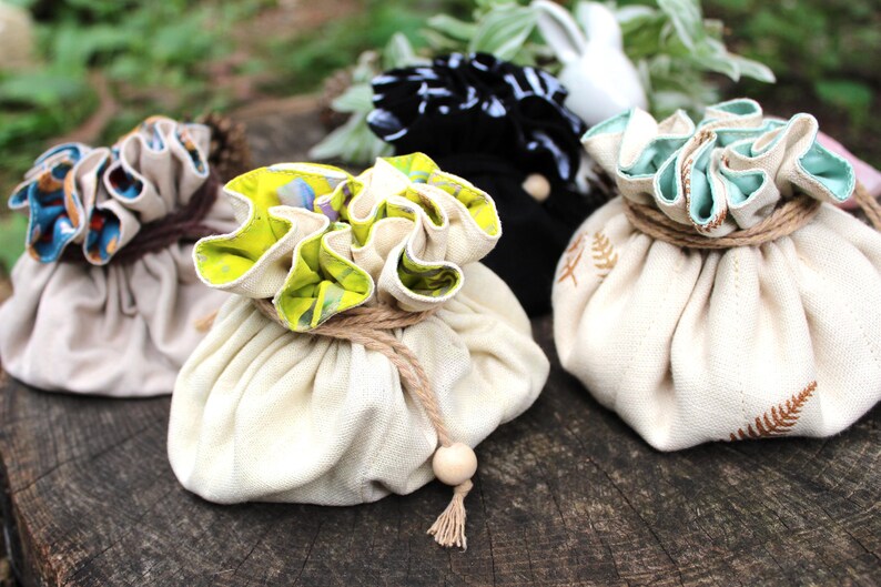 Puede incluir: Una colecci&oacute;n de bolsas de tela con cord&oacute;n en varios colores y patrones. Las bolsas est&aacute;n atadas con hilo y presentan bordes con volantes decorativos. Algunas tienen dise&ntilde;os florales o de helechos, y un detalle de cuentas de madera.