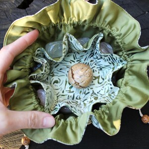 May include: A green and white patterned pouch with a drawstring closure, filled with various stones and a round, textured stone in the centre. The pouch is held open, showcasing the contents. Decorative beads and crystals are visible.