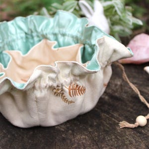 Puede incluir: Una bolsa con cord&oacute;n de color crema con forro de sat&eacute;n verde menta e interior beige. La bolsa presenta un borde con volantes y detalles de hojas marrones bordadas. Una cuenta de madera y una borla acent&uacute;an el cord&oacute;n.
