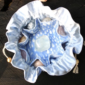 Könnte beinhalten: Ein hellblauer Satin-Kordelzugbeutel mit gerüschtem Rand und einem hellblauen Innenfutter mit weißem Regentropfenmuster. Der Beutel enthält mehrere kleine, glatte, polierte Steine in verschiedenen Farben, darunter ein klarer Kristall in der Mitte.