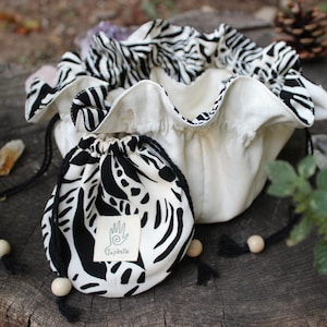 May include: Two drawstring bags with a black and white zebra print pattern. One bag is small and round, the other is a larger, ruffled bowl shape. Both have black drawstrings with wooden beads. The small bag has a white label with a hand symbol.