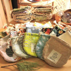 May include: A collection of small fabric drawstring pouches in various colors and patterns, including solid beige, lime green with yellow stripes, and floral prints. Each pouch features a white square label with a hand icon and the text Jujibelle.