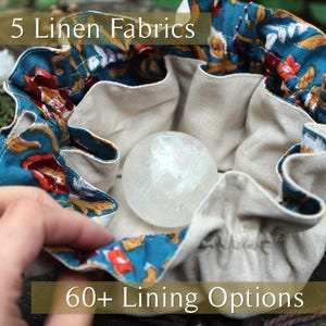 Puede incluir: Una bolsa de lino beige con cord&oacute;n y ribete con estampado floral turquesa, que contiene una bola de cristal transparente. La bolsa tiene el texto "5 Linen Fabrics" y "60+ Lining Options".