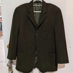 May include: A dark olive green blazer featuring a notched lapel, three front buttons, one chest pocket, and two lower patch pockets. The jacket is fully lined with a matching olive green fabric and is displayed on a light grey clothes hanger.