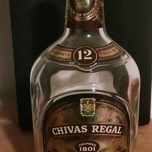 Op de afbeelding: Een heldere glazen fles Chivas Regal 12 jaar oude blended Scotch whisky. De fles heeft een gouden etiket met het Chivas Regal logo en de woorden "Gevestigd in 1801" en "Blended Scotch Whisky". De fles staat op een houten oppervlak.