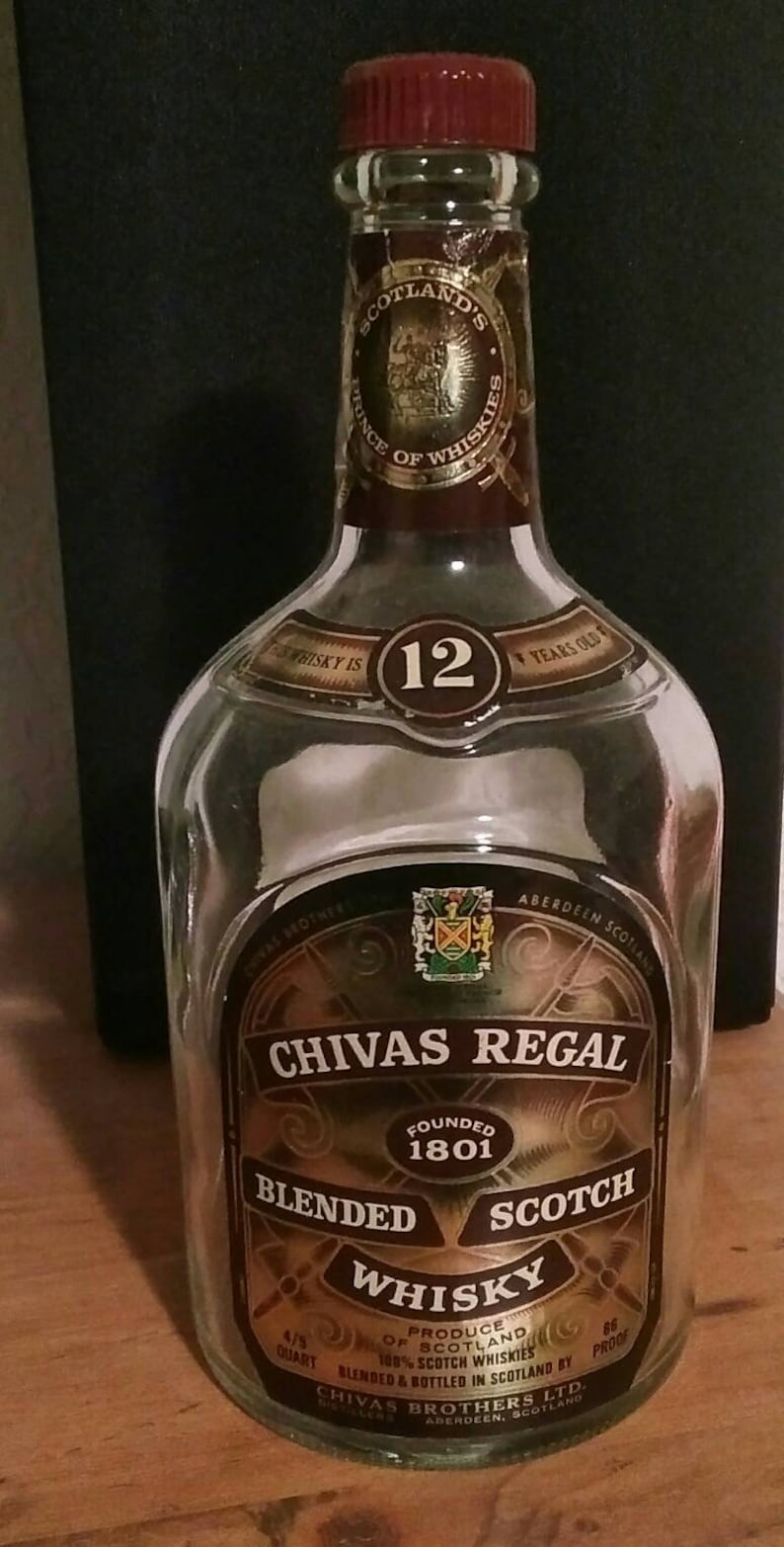 Op de afbeelding: Een heldere glazen fles Chivas Regal 12 jaar oude blended Scotch whisky. De fles heeft een gouden etiket met het Chivas Regal logo en de woorden "Gevestigd in 1801" en "Blended Scotch Whisky". De fles staat op een houten oppervlak.