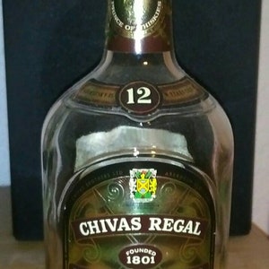 Op de afbeelding: Een heldere glazen fles Chivas Regal 12 jaar oude blended Scotch whisky. De fles heeft een rode dop en een gouden etiket met het Chivas Regal logo en de woorden "Gevestigd in 1801".