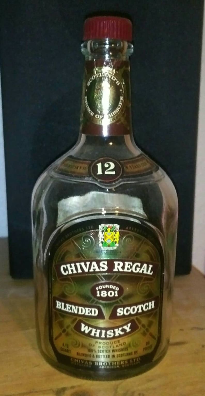 Op de afbeelding: Een heldere glazen fles Chivas Regal 12 jaar oude blended Scotch whisky. De fles heeft een rode dop en een gouden etiket met het Chivas Regal logo en de woorden "Gevestigd in 1801".