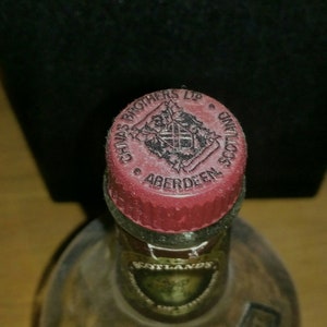 Op de afbeelding: Close-up van een rode dop met een zwart-wit logo met de tekst "Chivas Brothers Ltd. Aberdeen Scotland".
