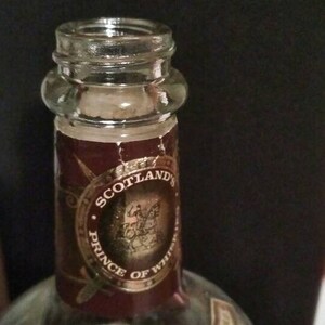 Op de afbeelding: Een doorzichtige glazen fles met een bruin etiket waarop "Scotland's Prince of Whiskies" staat en een gouden cirkel met het nummer 12. Het etiket heeft ook een gouden ontwerp van een ridder te paard.
