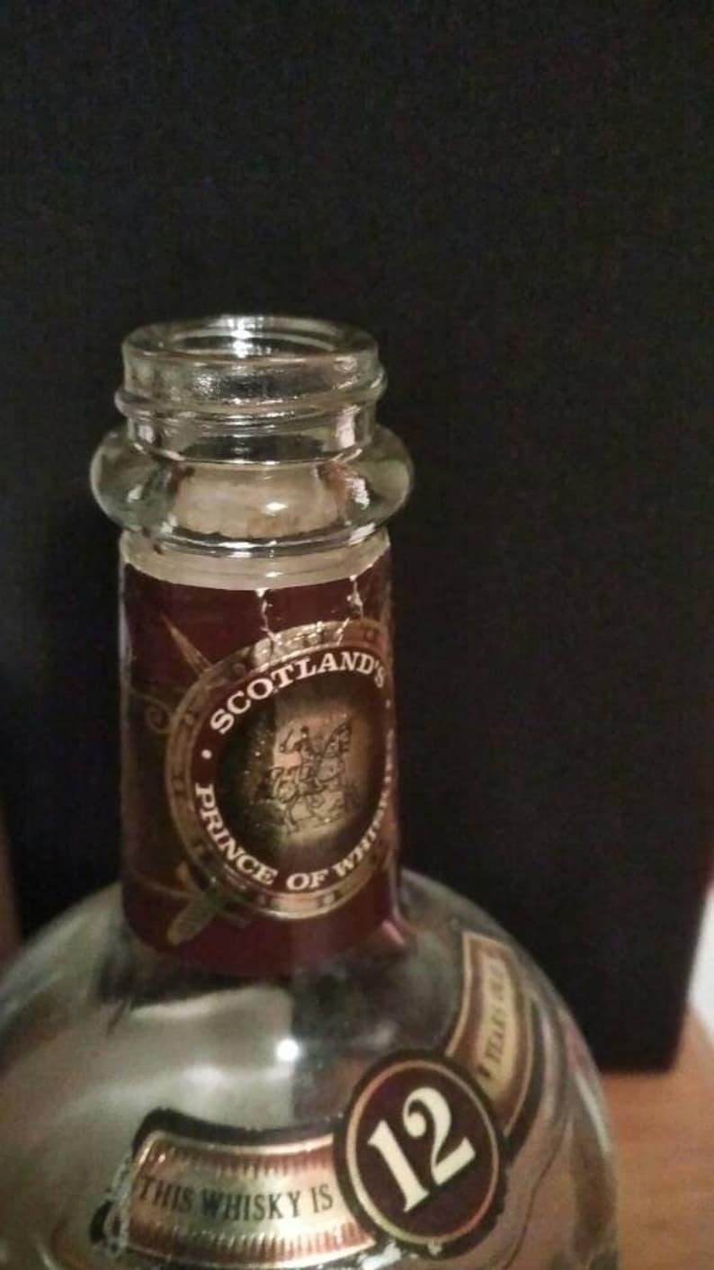 Op de afbeelding: Een doorzichtige glazen fles met een bruin etiket waarop "Scotland's Prince of Whiskies" staat en een gouden cirkel met het nummer 12. Het etiket heeft ook een gouden ontwerp van een ridder te paard.