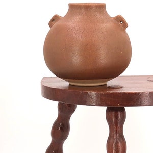 May include: A brown wooden stool with three legs. Each leg is made of a series of rounded knobs. A brown ceramic pot sits on top of the stool.