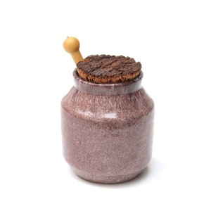 Puede incluir: Un pequeño tarro de cerámica moteada de color malva, con tapa de corcho y cuchara de madera. El tarro tiene un cuerpo redondeado y un cuello estrecho. La tapa de corcho es marrón oscuro y texturizada.