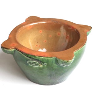 Può includere: Mortaio e pestello in ceramica verde con bordo marrone e macchie arancioni all'interno. Il mortaio ha il logo "KAVAL GK" sul lato.