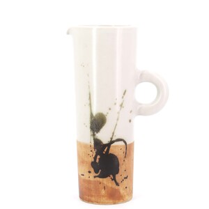 Puede incluir: Jarra alta de cerámica blanca con base marrón y asa. Presenta salpicaduras abstractas de pintura negra y verde. El asa es un simple bucle. La jarra se utiliza para servir bebidas. Altura aproximada de 20 cm.