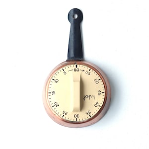 Peut inclure: Un minuteur de cuisine de couleur cuivre avec une poignée noire et un cadran blanc. Le minuteur comporte des chiffres de 5 à 60 et une flèche pointant vers l'heure actuelle. Le minuteur est réglé sur 60 minutes.