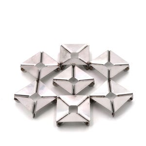May include: Eight silver-coloured, geometric trivets arranged in a flower-like pattern. Each trivet is square with a raised, pyramidal centre and small feet. The trivets are designed to protect surfaces from heat.
