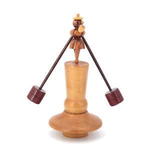May include: A wooden toy with a small figure in a hat and two wooden arms with weights on the ends. The figure is standing on a wooden base with a round top.