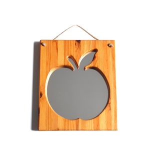 May include: A wooden mirror with an apple-shaped cutout. The mirror is framed in natural wood with a light brown finish. The mirror is hung with a simple rope.