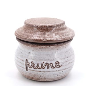 Peut inclure: Un pot en céramique blanche avec un couvercle brun. Le pot porte l'inscription "kruime" sur le côté.