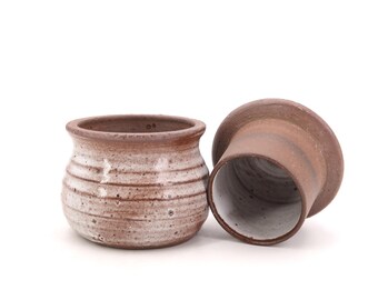 Può includere: Un set di due portaburro in ceramica marrone e bianca. I portaburro sono smaltati e hanno un aspetto rustico. Il portaburro superiore è un coperchio per il portaburro inferiore.