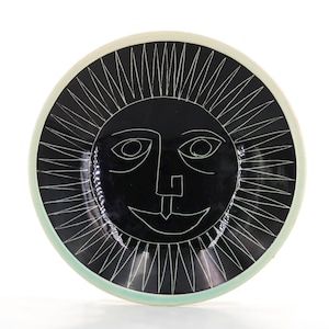 Puede incluir: Un plato redondo de cerámica con un esmalte negro brillante que presenta un rostro de sol dibujado con líneas blancas en el centro. El sol tiene ojos estilizados en forma de almendra, una nariz geométrica y una boca curva, rodeado de rayos puntiagudos.